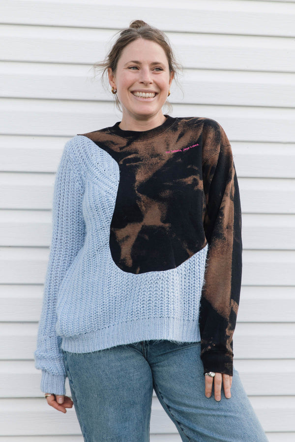 A woman smiling while wearing a color-blocked knit sweater with a light blue left side, a black right side, and a large brown tie-dye chest panel, paired with blue jeans, standing against a white siding background.