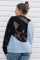 A woman shown from the back wearing a color-block knit sweater with a black and brown abstract pattern on the upper back and sleeves, a light blue chunky-knit lower body, and blue jeans against a white siding background.