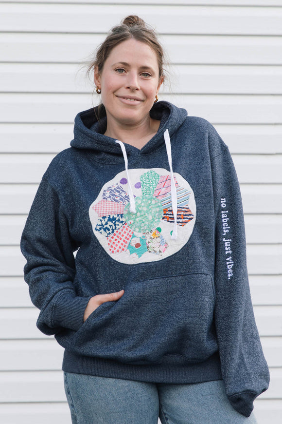 A woman wearing a navy blue heathered hoodie with a colorful circular patchwork graphic on the chest and white drawstrings, standing against a white siding wall.