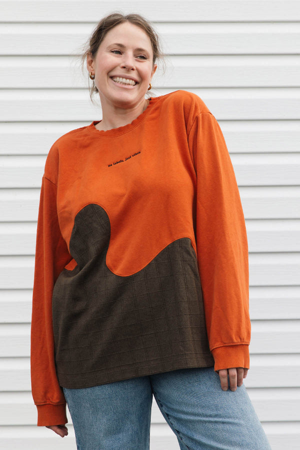 A woman wearing a long-sleeve color-block sweatshirt in burnt orange and dark brown, paired with blue jeans, standing against a white horizontal siding wall.