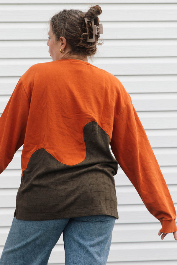 A woman shown from the back wearing a long-sleeve color-block top with a bright orange upper section and a dark brown lower section, paired with blue jeans against a white siding background.