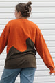 A woman shown from the back wearing a long-sleeve color-block top with a bright orange upper section and a dark brown lower section, paired with blue jeans against a white siding background.
