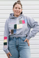 A woman wearing a light gray hoodie with colorful square patchwork accents on the chest and sleeve, paired with blue jeans, standing against a white siding background.