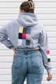 A woman viewed from behind wearing a light gray cropped hoodie with a large multicolor patchwork square on the back and matching patchwork details on the sleeve, paired with blue jeans against a white siding background.