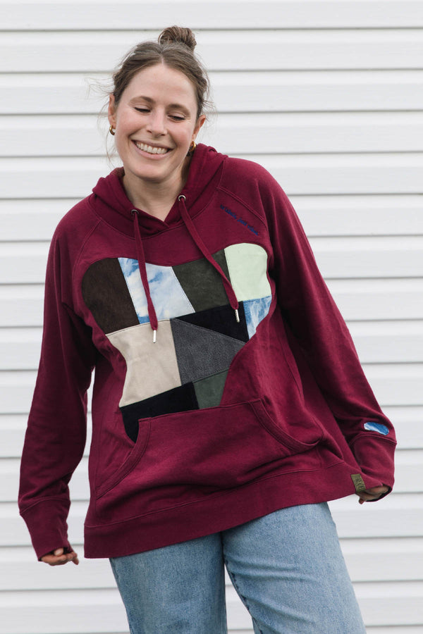 A woman wearing a burgundy pullover hoodie with a large multicolor geometric patchwork heart design on the front, paired with light blue jeans, standing against a white siding wall.
