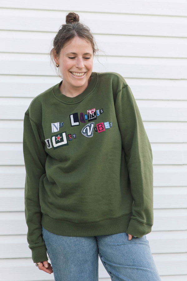 A woman wearing an olive green crewneck sweatshirt with colorful patchwork lettering on the front that reads 'no labels, just vibes.', paired with blue jeans, standing against a white horizontal siding wall.