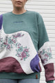 person wearing colourful flower sweater. Purple, Maroon and Green floral motif, upcycled in canada, detail shot featuring 'no labels, just vibes.' embroidery