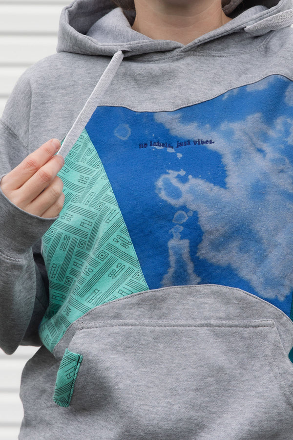person wearing grey hoodie with blue, teal and green colourful patches, upcycled in canada, detail shot featuring 'no label, just vibes.' embroidery