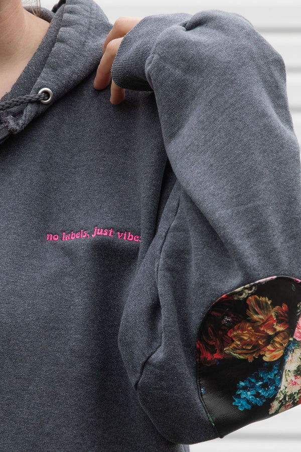 person wearing grey zip up hoodie with floral elbow patches, detail shot of 'no labels, just vibes.' embroidery