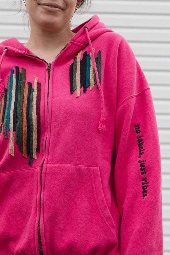 Person wearing a bright pink hoodie with a unique and colourful design, upcycled in canada, detail shot of 'no labels, just vibes.' embroidery