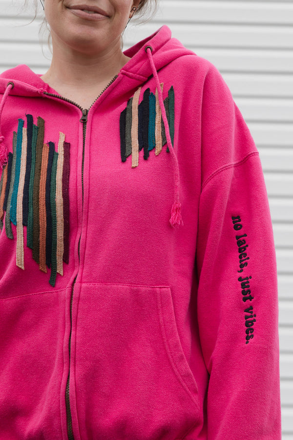 Person wearing a bright pink hoodie with a unique and colourful design, upcycled in canada, detail shot of 'no labels, just vibes.' embroidery