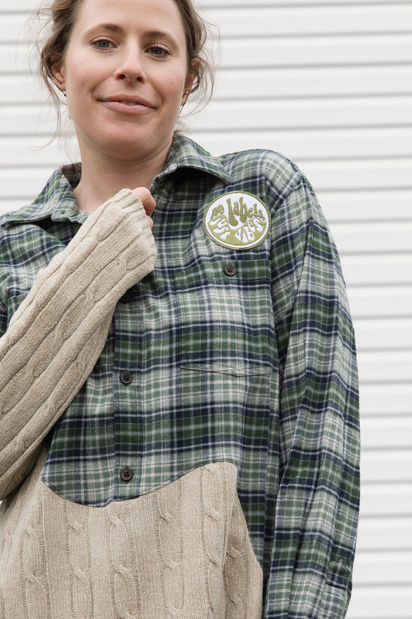 person wearing an upcycled shirt, green plaid and cream knit sewn together, repurposed in canada, detail shot of 'no labels, just vibes.' patch