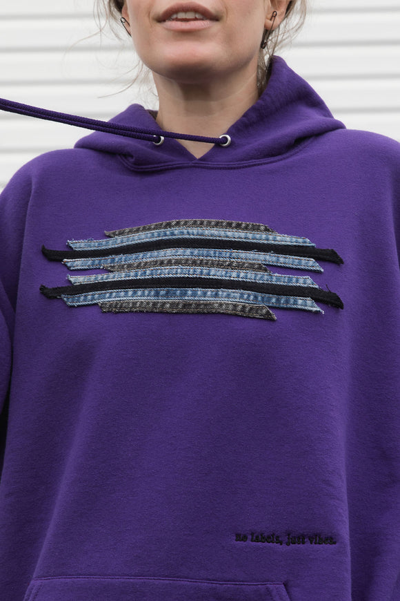 Person wearing a purple hoodie with a graphic design featuring denim seams, upcycled in canada, detail shot of 'no labels, just vibes.' embroidery