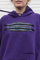 Person wearing a purple hoodie with a graphic design featuring denim seams, upcycled in canada, detail shot of 'no labels, just vibes.' embroidery