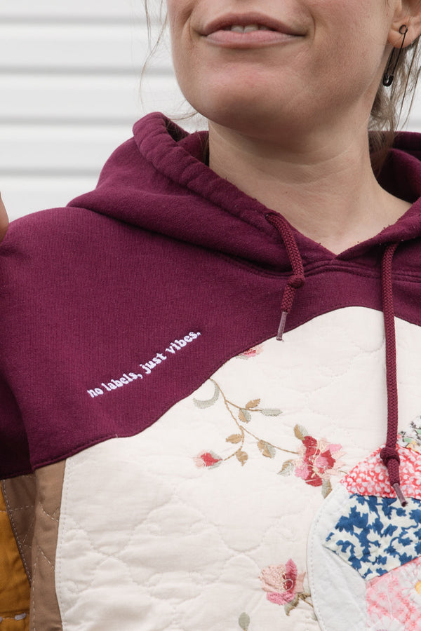 Person wearing a maroon hoodie with a colorful abstract design made with quilting scraps, upcycled in canada. detail shot of 'no labels, just vibes.' embroidery