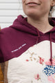 Person wearing a maroon hoodie with a colorful abstract design made with quilting scraps, upcycled in canada. detail shot of 'no labels, just vibes.' embroidery