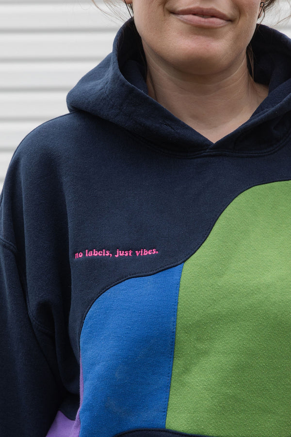 Person wearing a navy hoodie with a colourful rainbow design upcycled in canada detail shot of 'no labels, just vibes.' embroidery