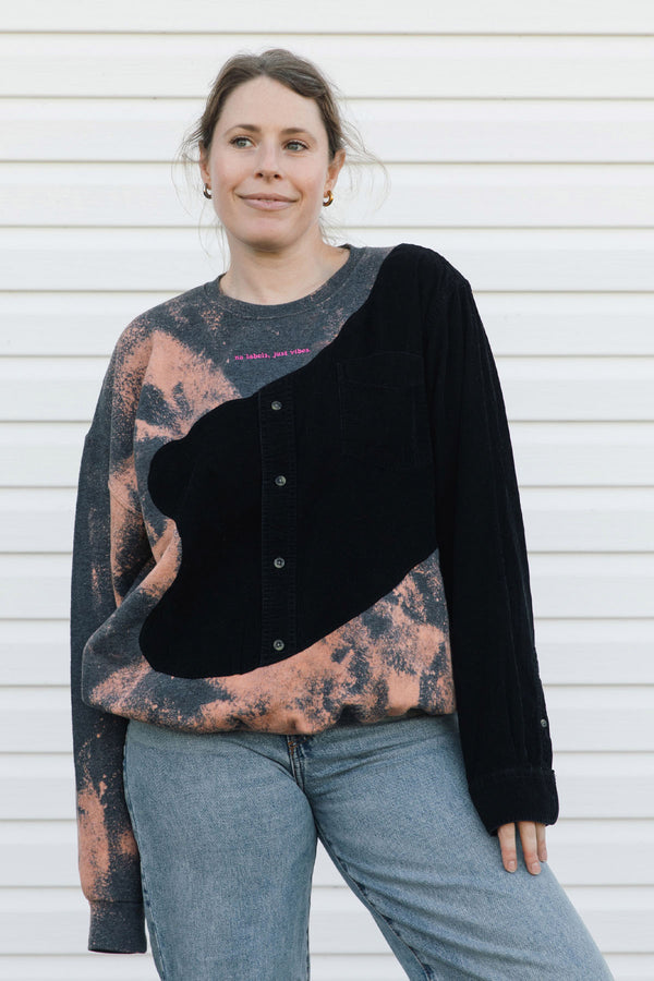 A woman wearing a unique asymmetrical cardigan sweater with black velvet and a pink-brown acid-wash pattern, paired with blue jeans, standing against a white siding background.