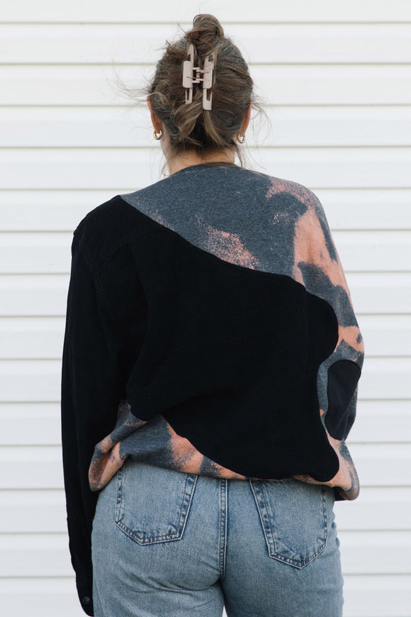 A woman viewed from behind wearing a long-sleeve knit sweater with a diagonal color-block design in black and a gray, pink, and peach tie-dye pattern, paired with light-wash blue jeans against a white siding background.