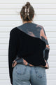 A woman viewed from behind wearing a long-sleeve knit sweater with a diagonal color-block design in black and a gray, pink, and peach tie-dye pattern, paired with light-wash blue jeans against a white siding background.