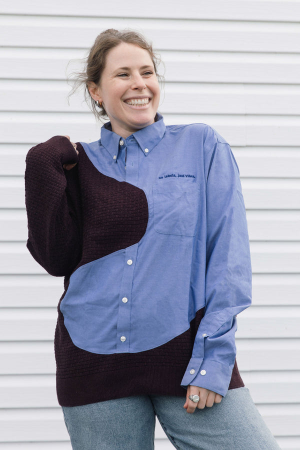 A woman smiling and wearing a color-block button-down shirt with a light blue upper section, dark burgundy lower section, and matching dark burgundy sleeves, paired with blue jeans in front of a white siding background.