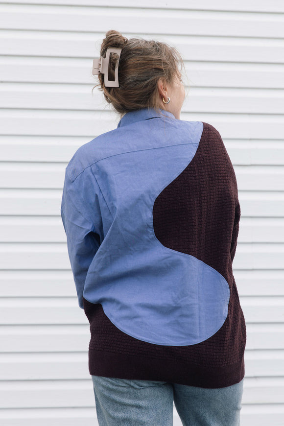 A woman smiling and wearing a color-block button-down shirt with a light blue upper section, dark burgundy lower section, and matching dark burgundy sleeves, paired with blue jeans in front of a white siding background.