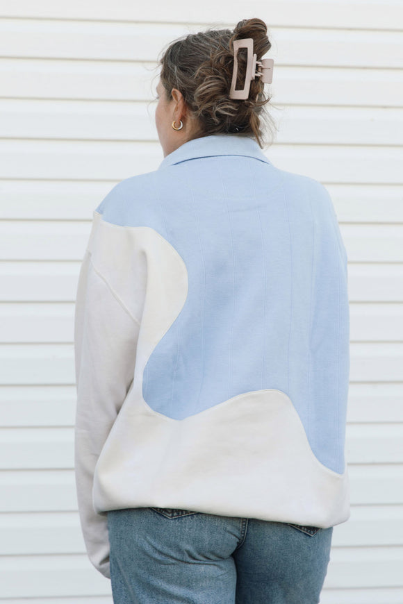 A woman viewed from behind wearing a color-blocked  sweater with light blue and cream panels, paired with blue jeans against a white siding background.