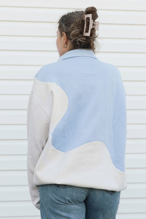 A woman viewed from behind wearing a color-blocked  sweater with light blue and cream panels, paired with blue jeans against a white siding background.