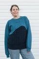 A woman wearing a color-block crewneck sweater with a teal blue upper section and a dark navy cable-knit lower section, paired with light blue jeans, standing against a white horizontal siding wall.
