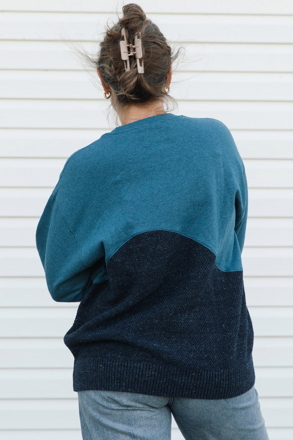 A person viewed from behind wearing a color-block knit sweater with a teal upper section and a dark navy lower section, paired with light blue jeans against a white siding background.