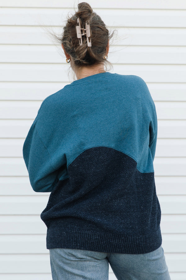 A person viewed from behind wearing a color-block knit sweater with a teal upper section and a dark navy lower section, paired with light blue jeans against a white siding background.