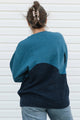 A person viewed from behind wearing a color-block knit sweater with a teal upper section and a dark navy lower section, paired with light blue jeans against a white siding background.