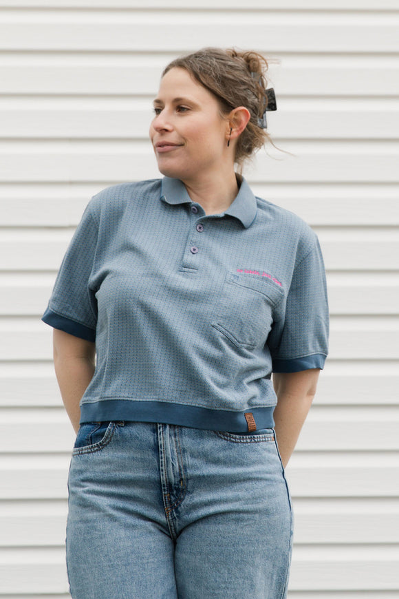 Woman wearing a blue polo shirt with a logo and jeans against a white wall.