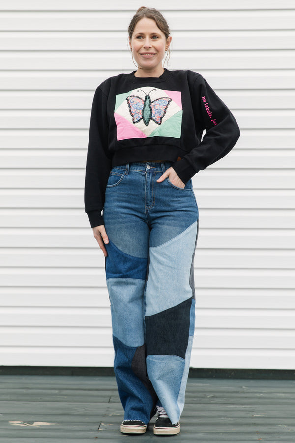 Woman wearing a black sweater with a colorful butterfly design and patchwork jeans against a white background.