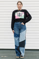 Woman wearing a black sweater with a colorful butterfly design and patchwork jeans against a white background.