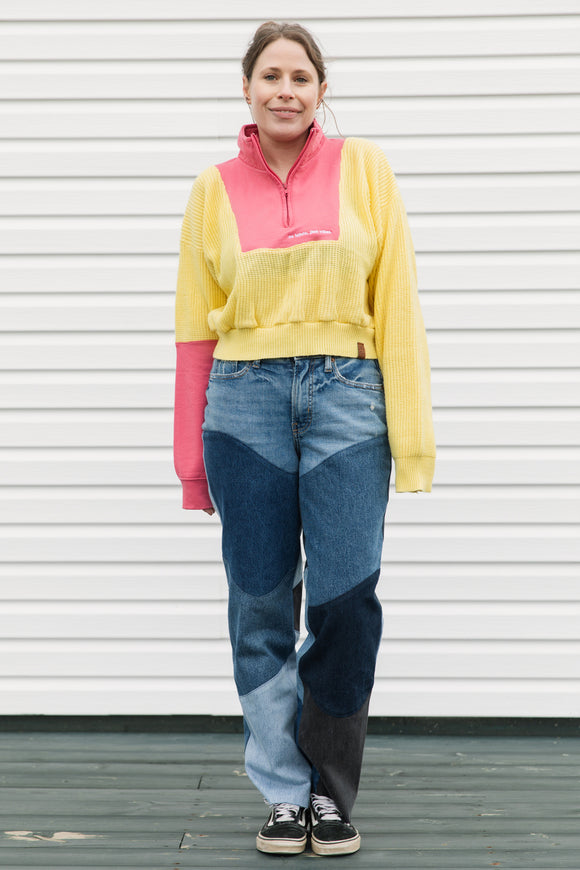 Person wearing a colorful sweater and patchwork jeans standing against a white wall.