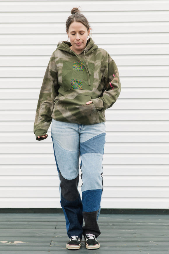 Person wearing a green camouflage hoodie and patchwork jeans standing against a white wall.