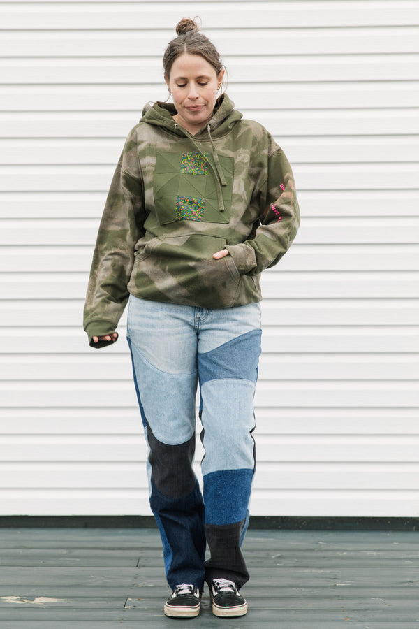 Person wearing a green camouflage hoodie and patchwork jeans standing against a white wall.