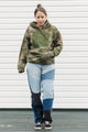 Person wearing a green camouflage hoodie and patchwork jeans standing against a white wall.