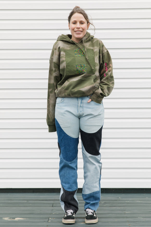 Person wearing a green hoodie with floral design and patchwork jeans standing against a white wall.