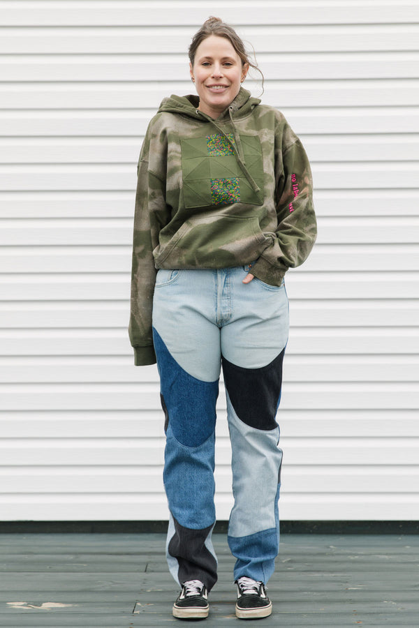 Person wearing a green hoodie with floral design and patchwork jeans standing against a white wall.
