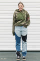 Person wearing a green hoodie with floral design and patchwork jeans standing against a white wall.