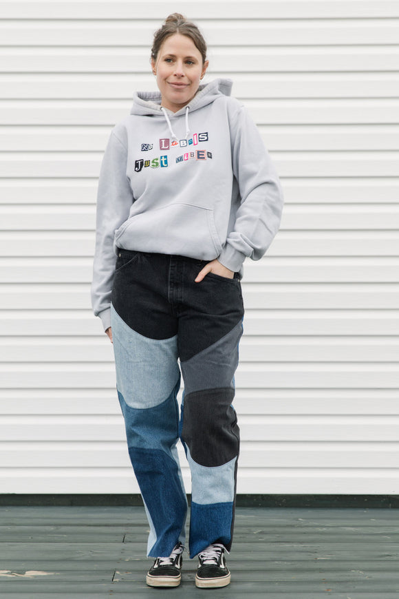 Person wearing a gray hoodie with colorful text and black and blue patchwork pants against a white wall.