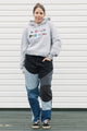Person wearing a gray hoodie with colorful text and black and blue patchwork pants against a white wall.