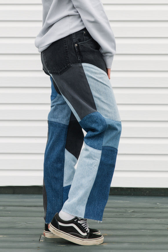 Person wearing a pair of patchwork jeans with black, blue, and gray colors against a white wall.