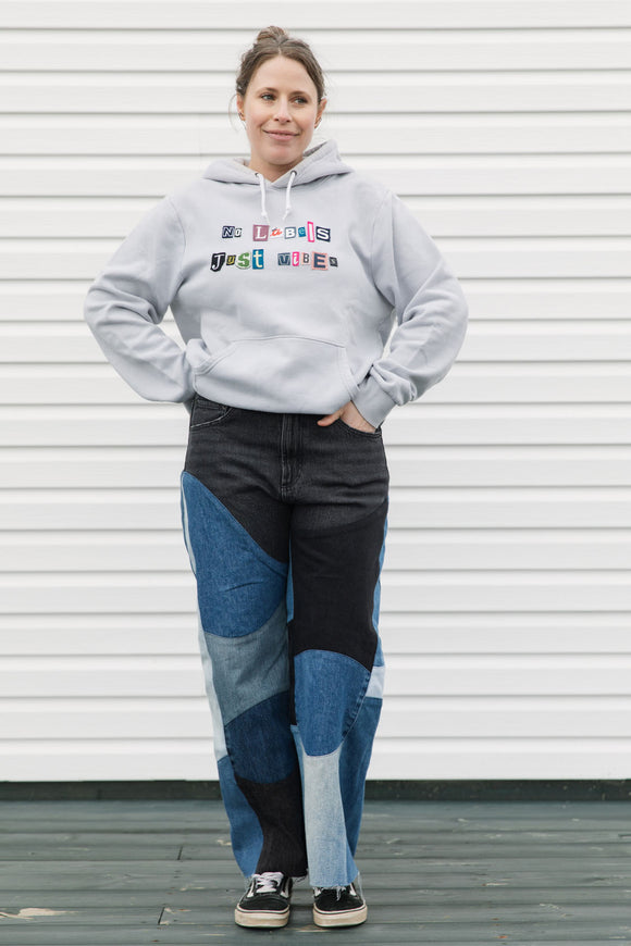 Person wearing a gray hoodie with colorful text and patchwork jeans standing against a white wall, upcycled in canada