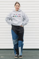 Person wearing a gray hoodie with colorful text and patchwork jeans standing against a white wall, upcycled in canada
