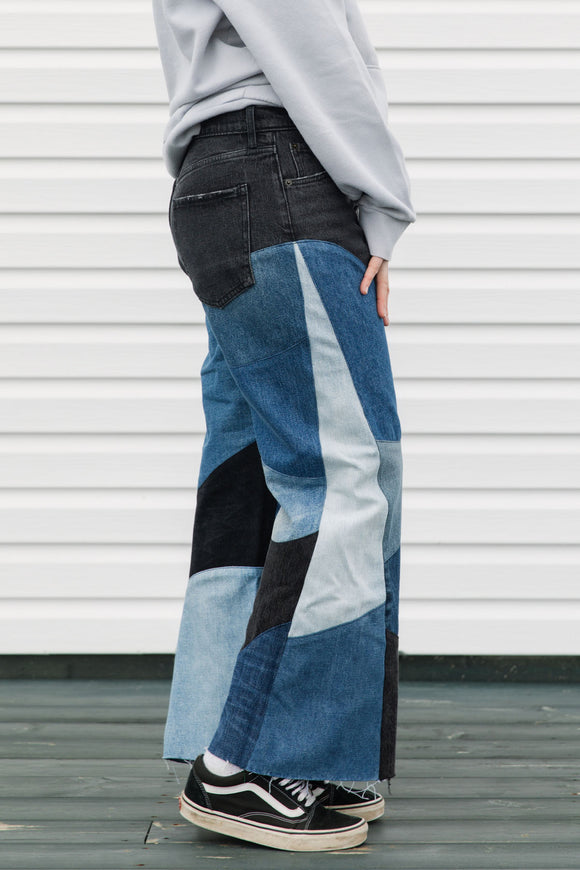 Person wearing a gray hoodie with colorful text and patchwork jeans standing against a white wall, upcycled in canada