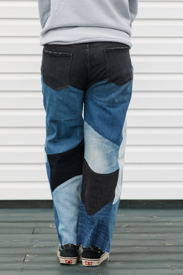 Person wearing a gray hoodie with colorful text and patchwork jeans standing against a white wall, upcycled in canada