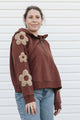 A woman wearing a brown cropped pullover hoodie with beige flower appliqués on the sleeves, paired with blue jeans, standing against a white siding background.